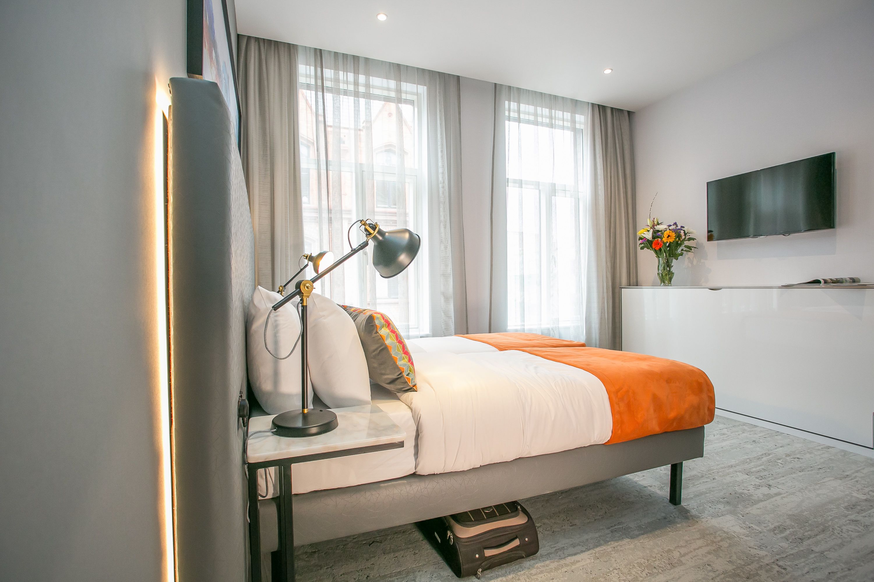 Una camera d'albergo moderna con un letto, una lampada da comodino, tende, una TV e un vaso con fiori.