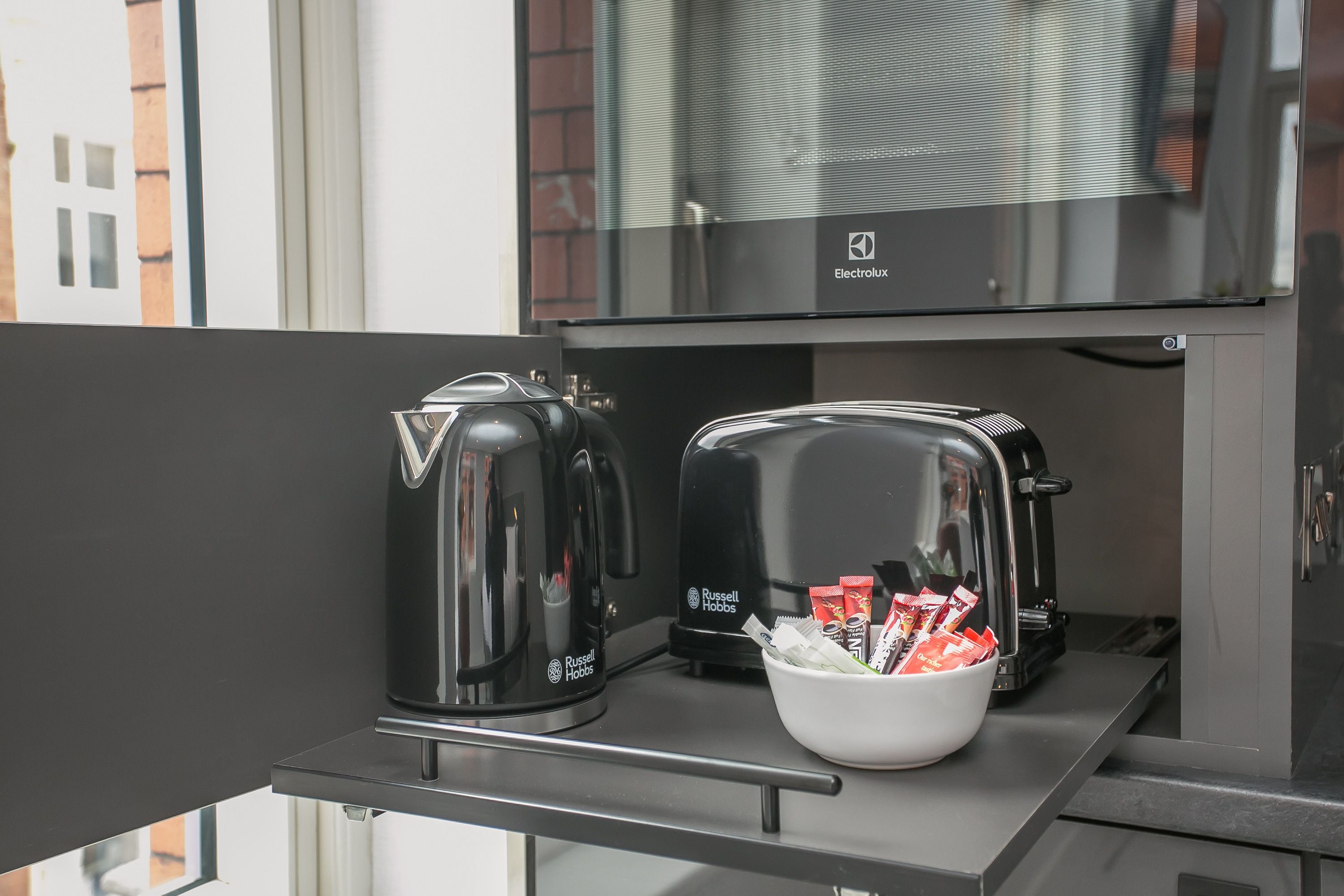 A kitchenette with two kettles, a toaster, and a bowl of snacks.