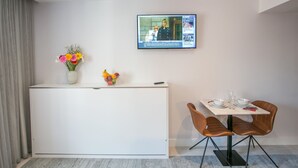 Una habitación de hotel moderna con un mostrador de recepción blanco, una zona de comedor con dos sillas marrones y una mesa, y un televisor de pantalla plana montado en la pared que muestra un noticiero.