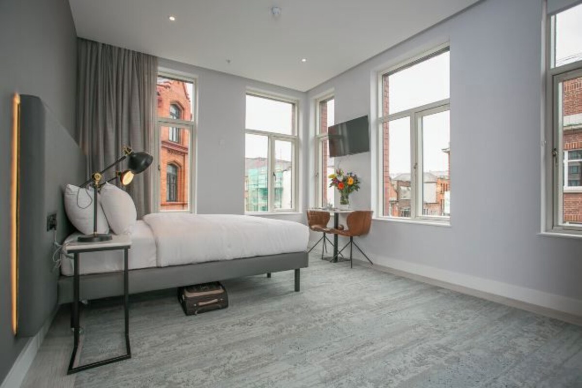 A modern bedroom with a bed, a desk, and a chair, large windows, and a TV on the wall.