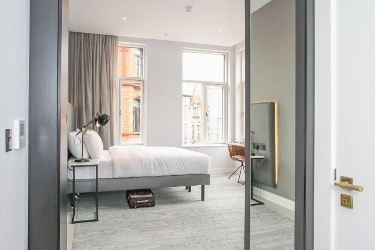A modern bedroom with a bed, bedside table, lamp, and a view of a building through a window.