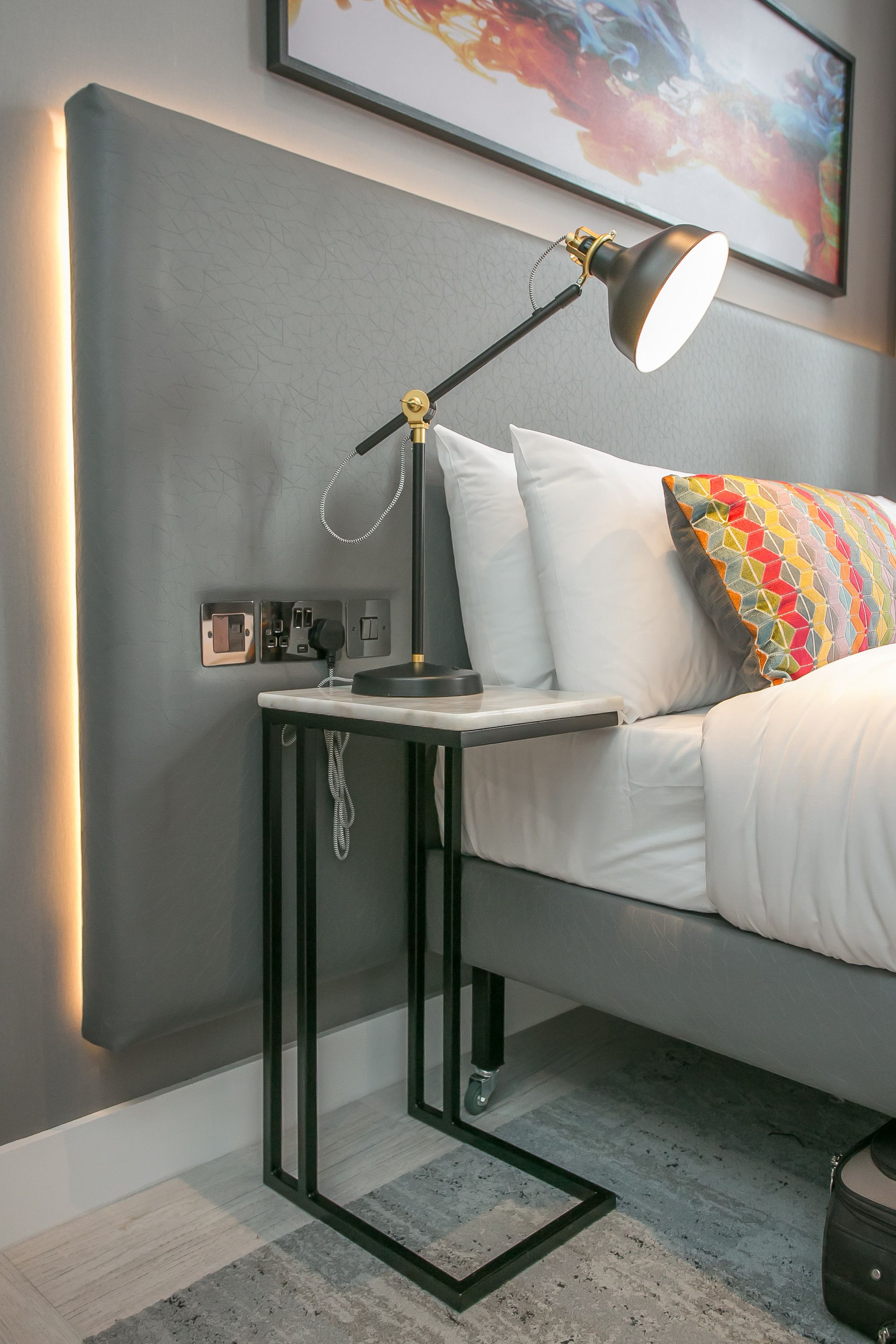 A modern hotel room with a bed, a bedside table with a lamp, a power outlet, and a wall-mounted switch.
