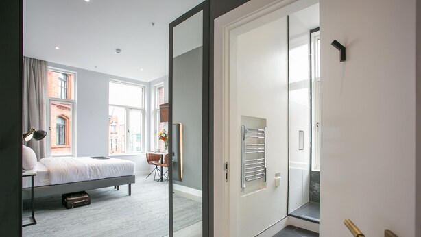Un dormitorio moderno con una cama, un escritorio y una silla. Hay un ventanal y una puerta que da a otra habitación.