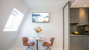 Habitación de hotel moderna con zona de comedor que incluye dos sillas marrones, una mesa pequeña con un jarrón de flores y un televisor de pantalla plana mostrando un mensaje de bienvenida.