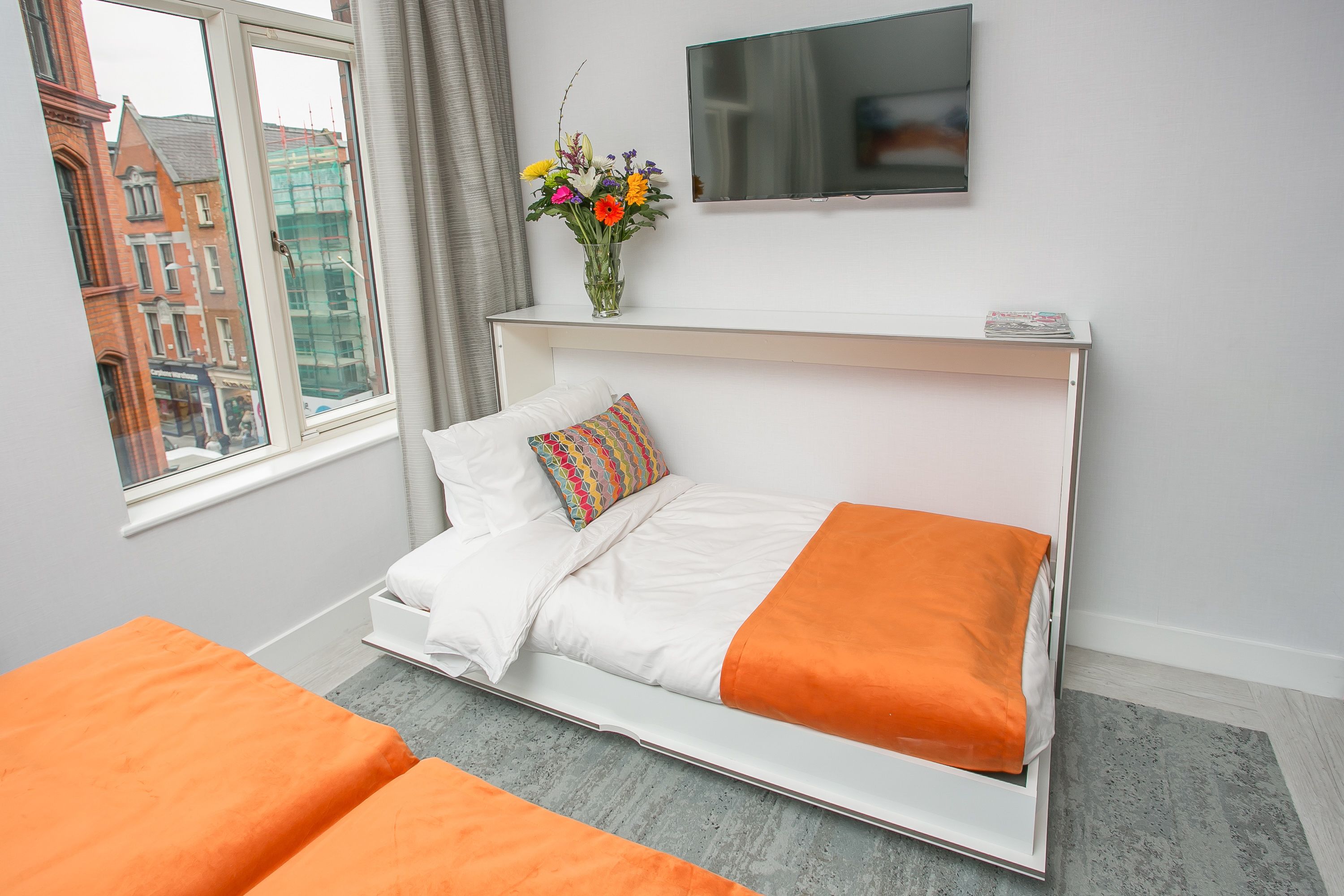 A modern hotel room with a bed, a TV, a vase with flowers, and a window with a city view.