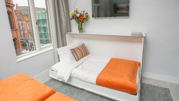 A modern hotel room with a bed, a TV, a vase with flowers, and a window with a city view.