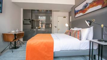 A modern hotel room with a bed, bedside table, lamp, and a view of the kitchen and dining area.