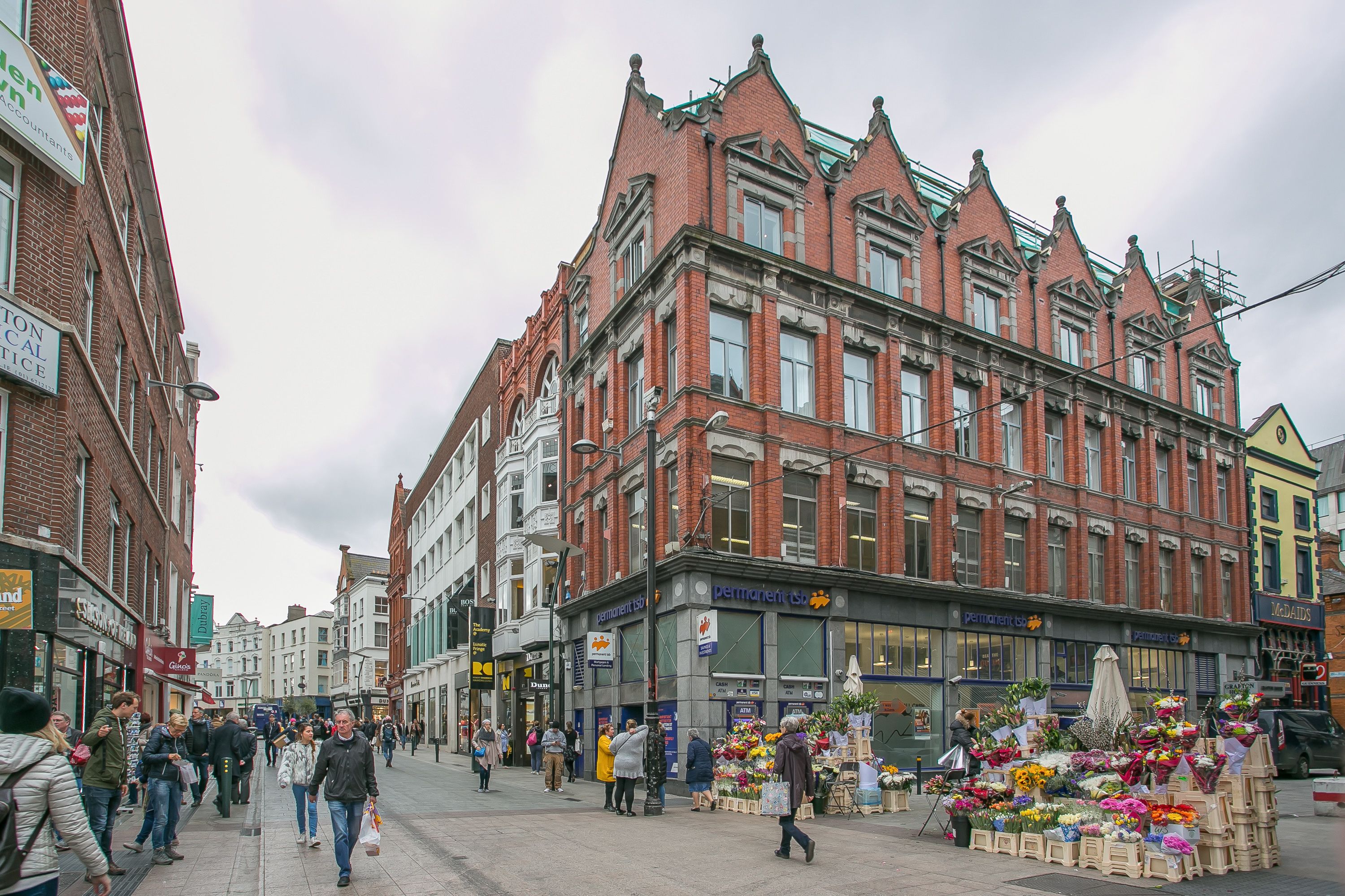A street with a row of red brick buildings, a flower market, and pedestrians.