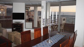A dining area with a table set for four, a television, and a view of the ocean through large windows.