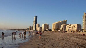 A beach with people walking along the shore, buildings in the background, and a clear sky.