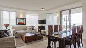A spacious living room with a sofa, coffee table, TV, and dining area with chairs and a chandelier.