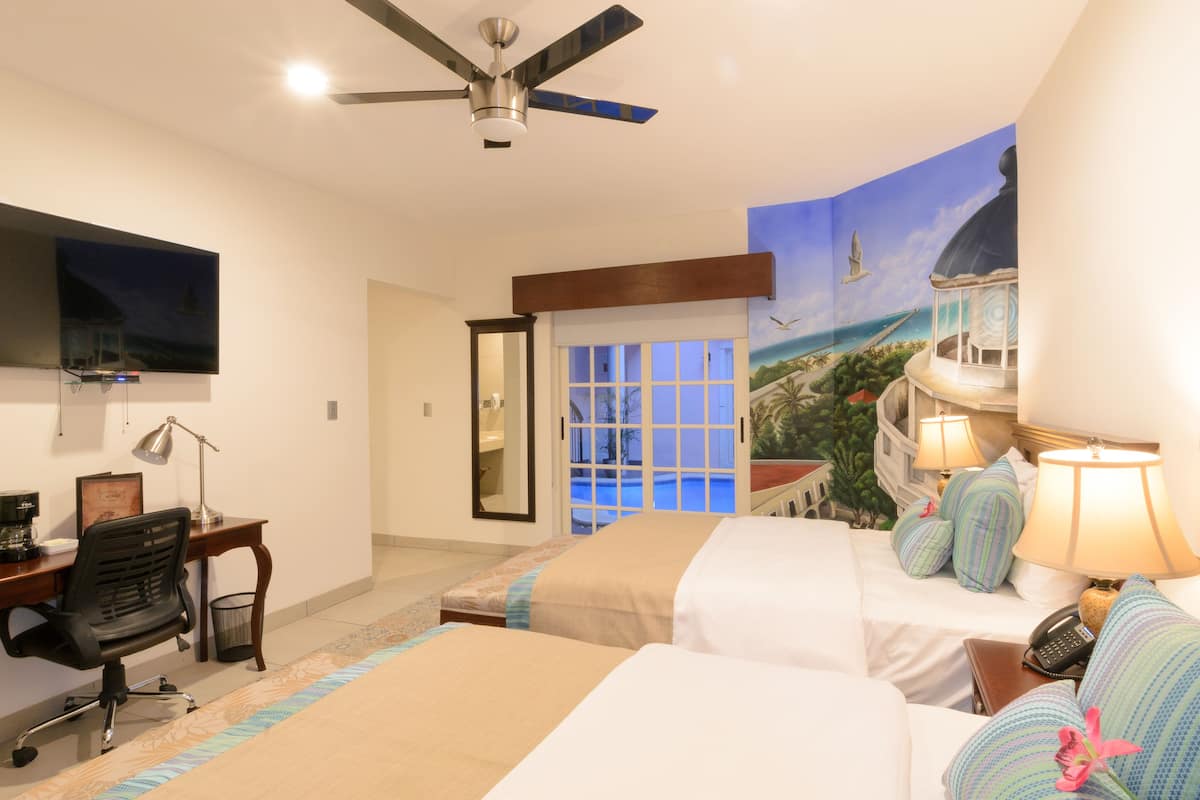 Habitación de hotel con dos camas, un escritorio con una silla, un televisor y un mural con una escena de playa.