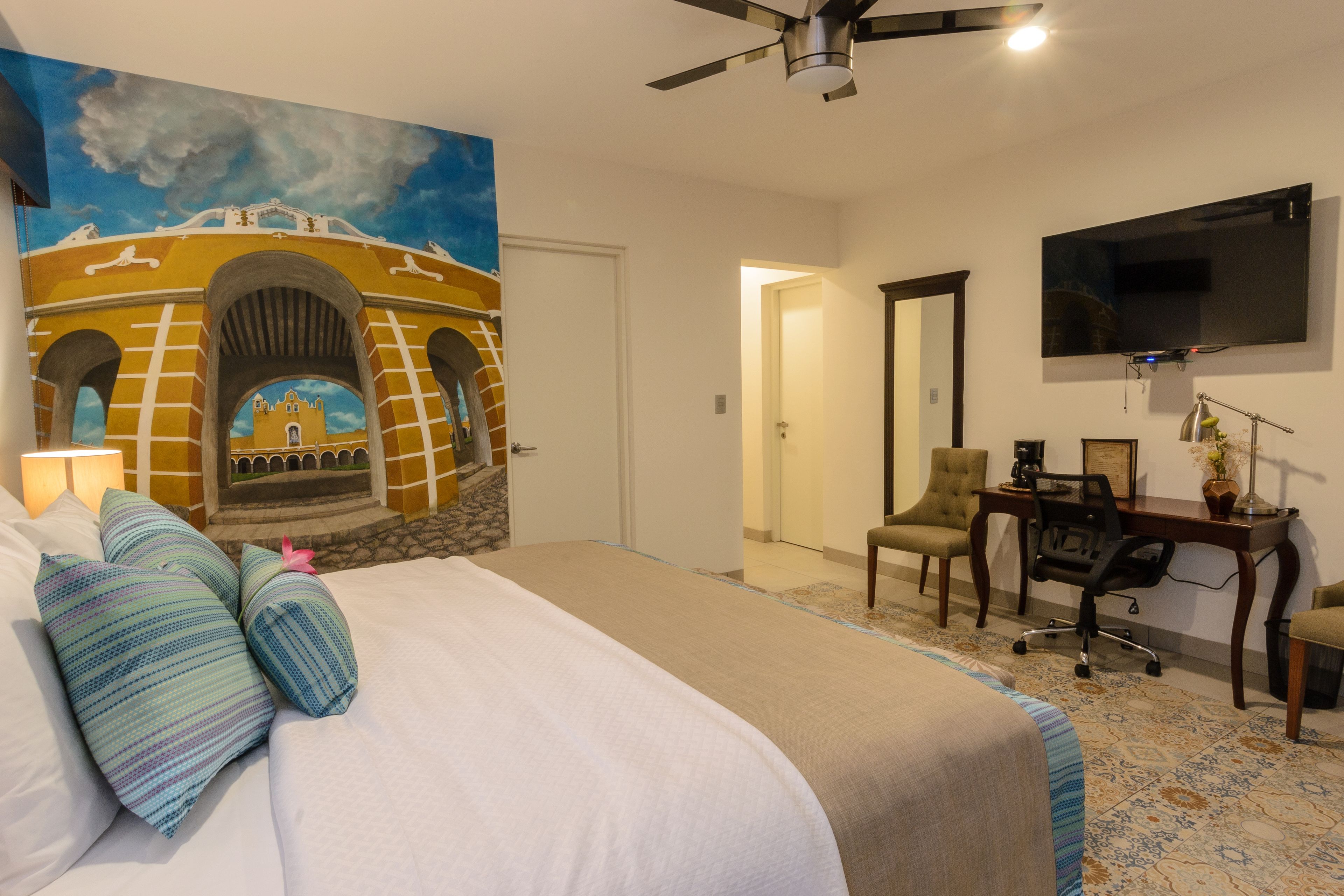 A hotel room with a bed, a desk, a chair, a TV, and a mural of a building.