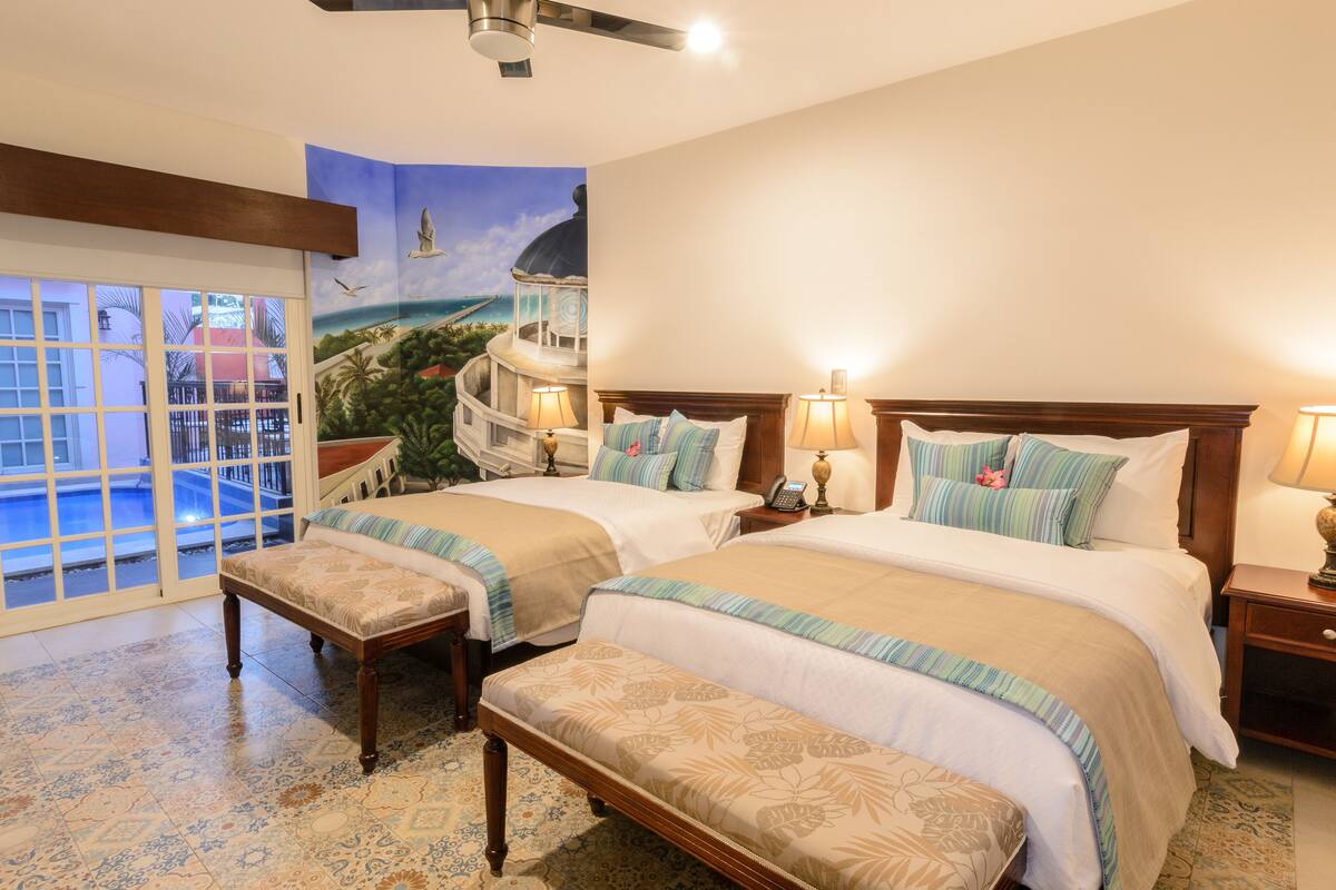 Habitación de hotel con dos camas, un balcón con piscina y un mural de paisaje tropical.