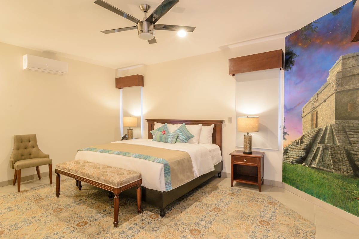 Habitación de hotel con cama, silla, banco, mesita de noche, ventilador de techo y un mural en la pared.