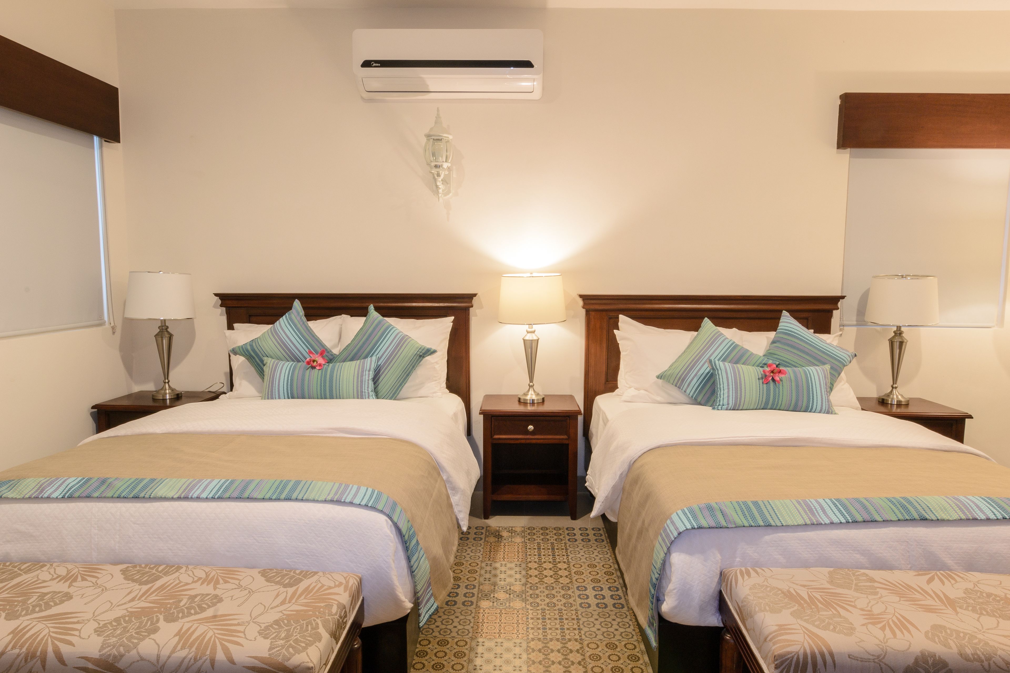 A hotel room with two beds, each with a wooden headboard, a bedside table with a lamp, and a patterned rug.