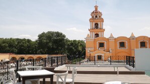 Una terraza en la azotea con mesas y sillas, con vistas a un edificio con una torre de campanas y ventanas de arco.