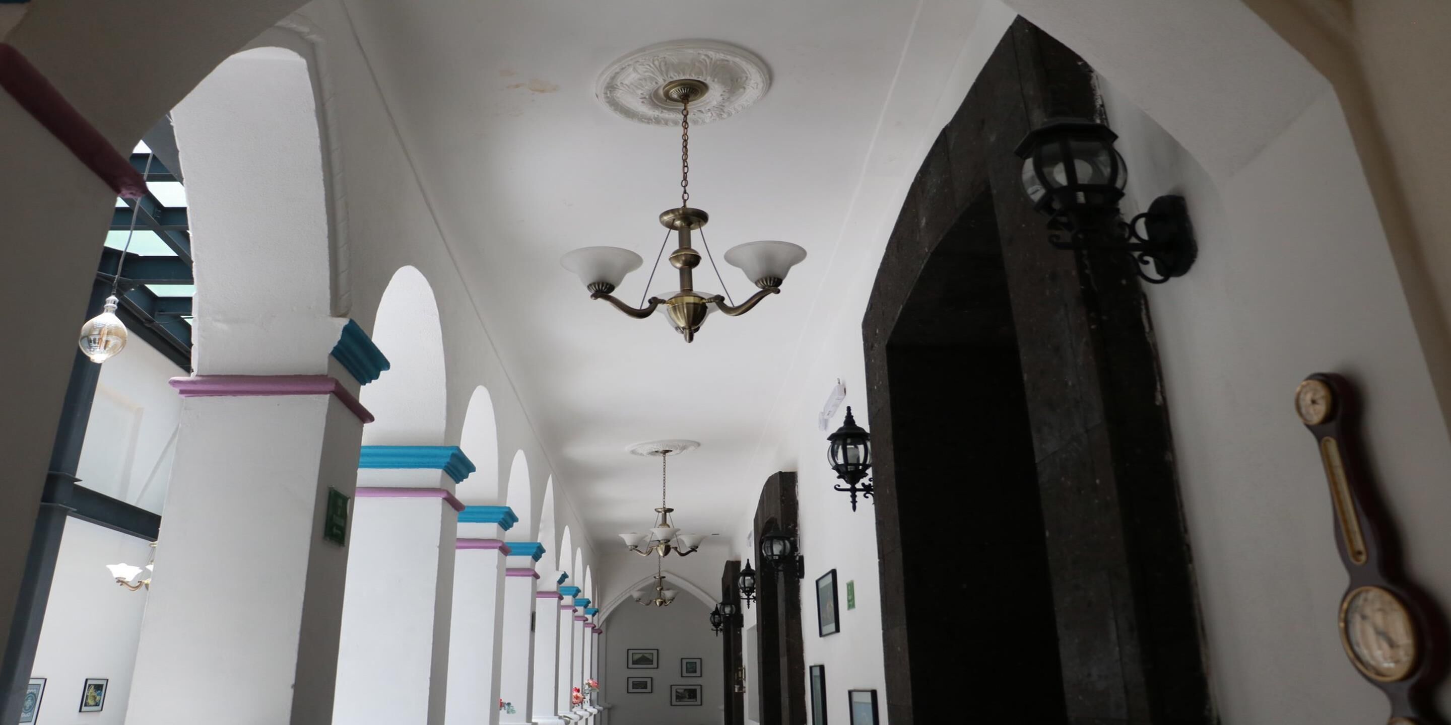 Un pasillo con paredes blancas, detalles en azul y rosa, candelabros y lámparas de pared.