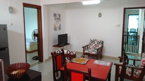 A dining area with a red table, wooden chairs, a television on a stand, and a decorative wall piece. - La Primerose (Quatre Bornes)