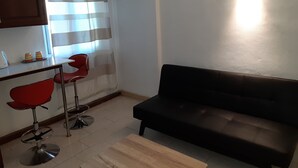 A living room with a black sofa, two red chairs, a wooden coffee table, a TV mounted on the wall, and a window with curtains. - La Primerose (Quatre Bornes)