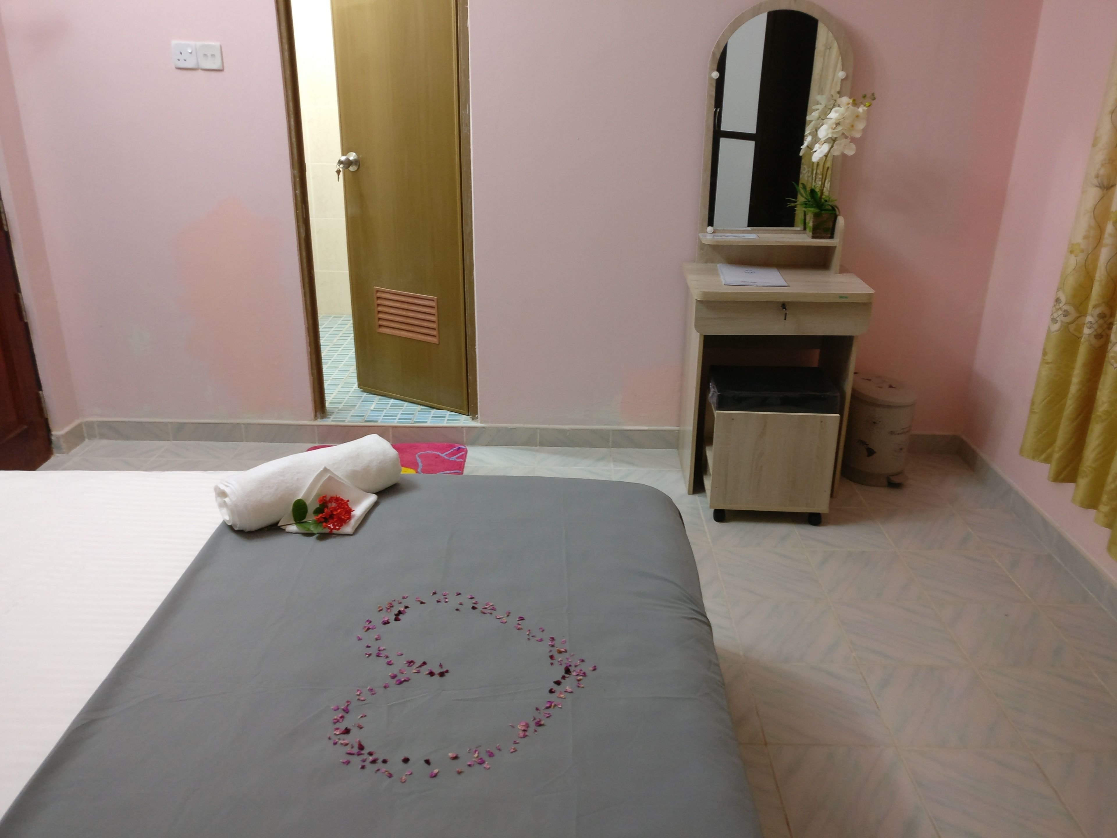 Una cama bien hecha con una toalla y un arreglo floral, un pequeño escritorio de madera con un espejo, un cubo de basura y una puerta que da a otra habitación.
