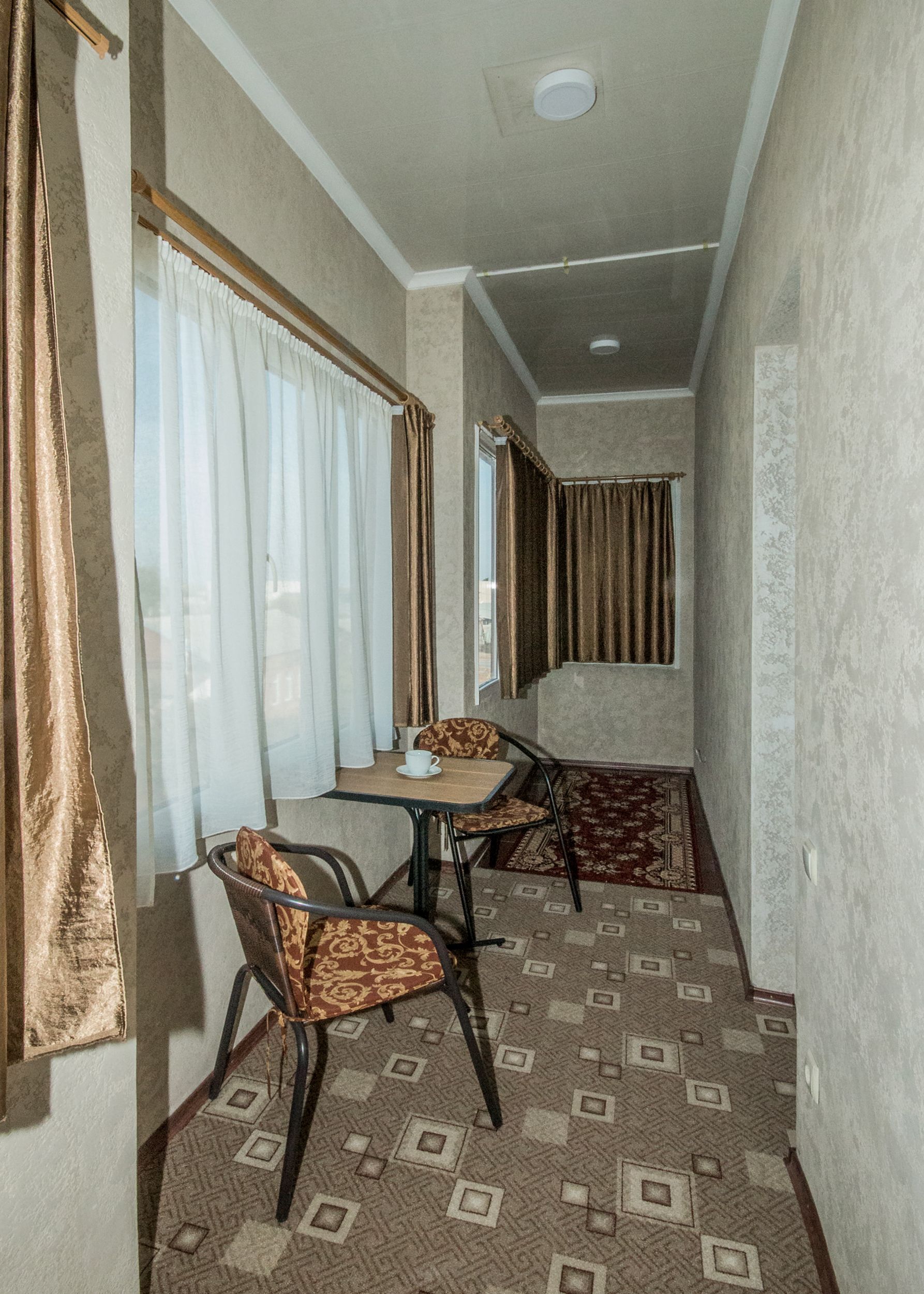 A narrow room with a small table and two chairs, a patterned carpet, and a window with curtains.