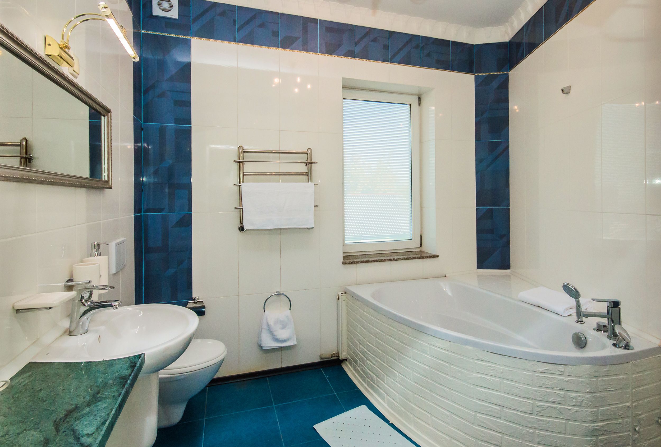 Un baño con lavamanos, inodoro y bañera blancos, azulejos azules y una ventana.