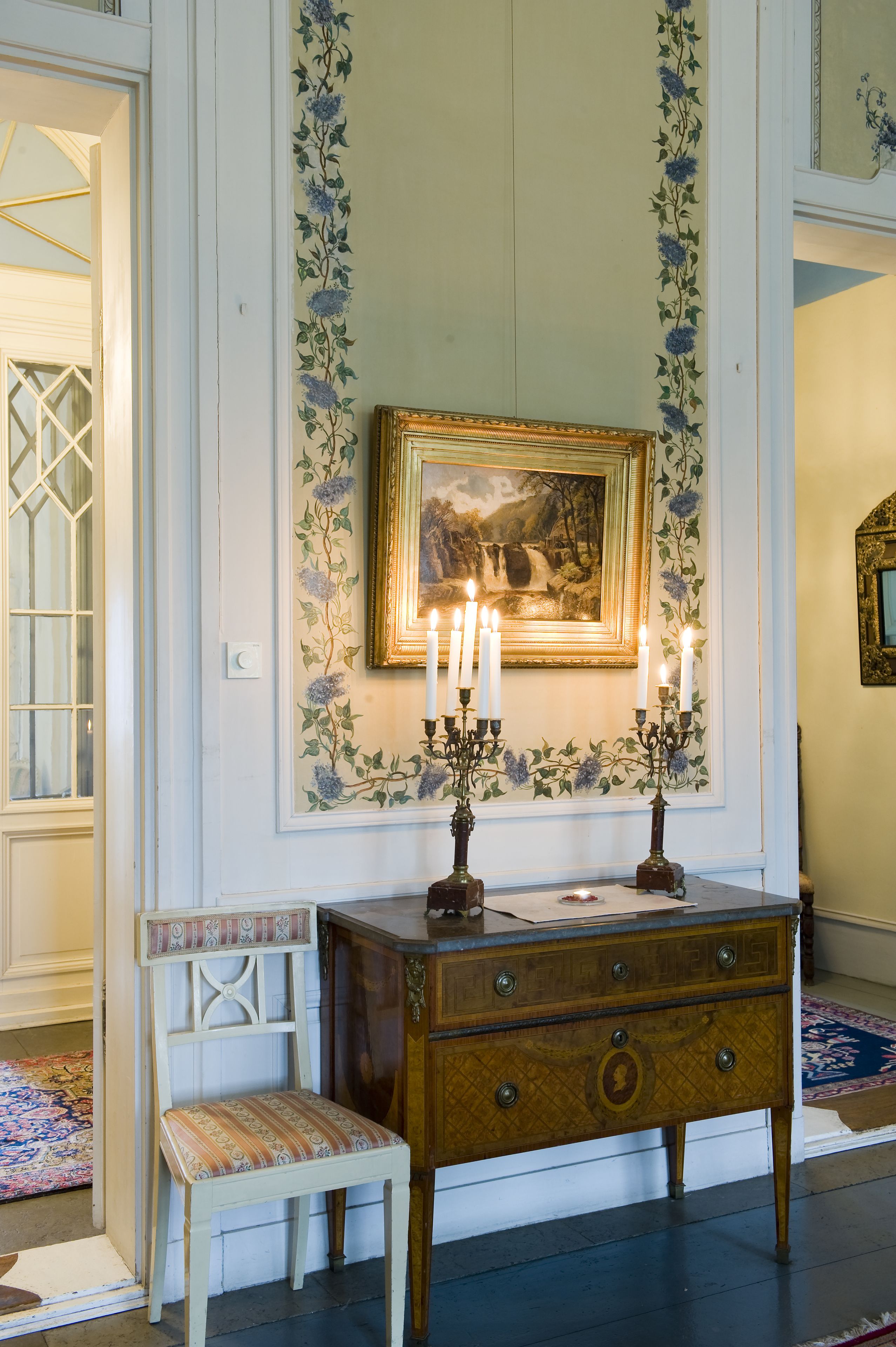 Une pièce comprenant une commode en bois, une chaise, un tableau encadré et un chandelier avec des bougies allumées.