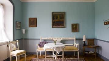 A room with a white bed, a round table with a white tablecloth, two chairs, a patterned rug, and framed pictures on the wall.