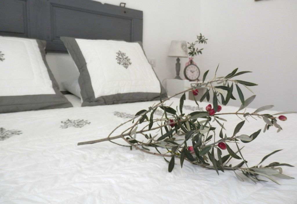 Een net opgemaakt bed met witte beddengoed en grijze kussens, een nachtkastje met een lamp en een klok, en een tak met rode bessen op het bed.