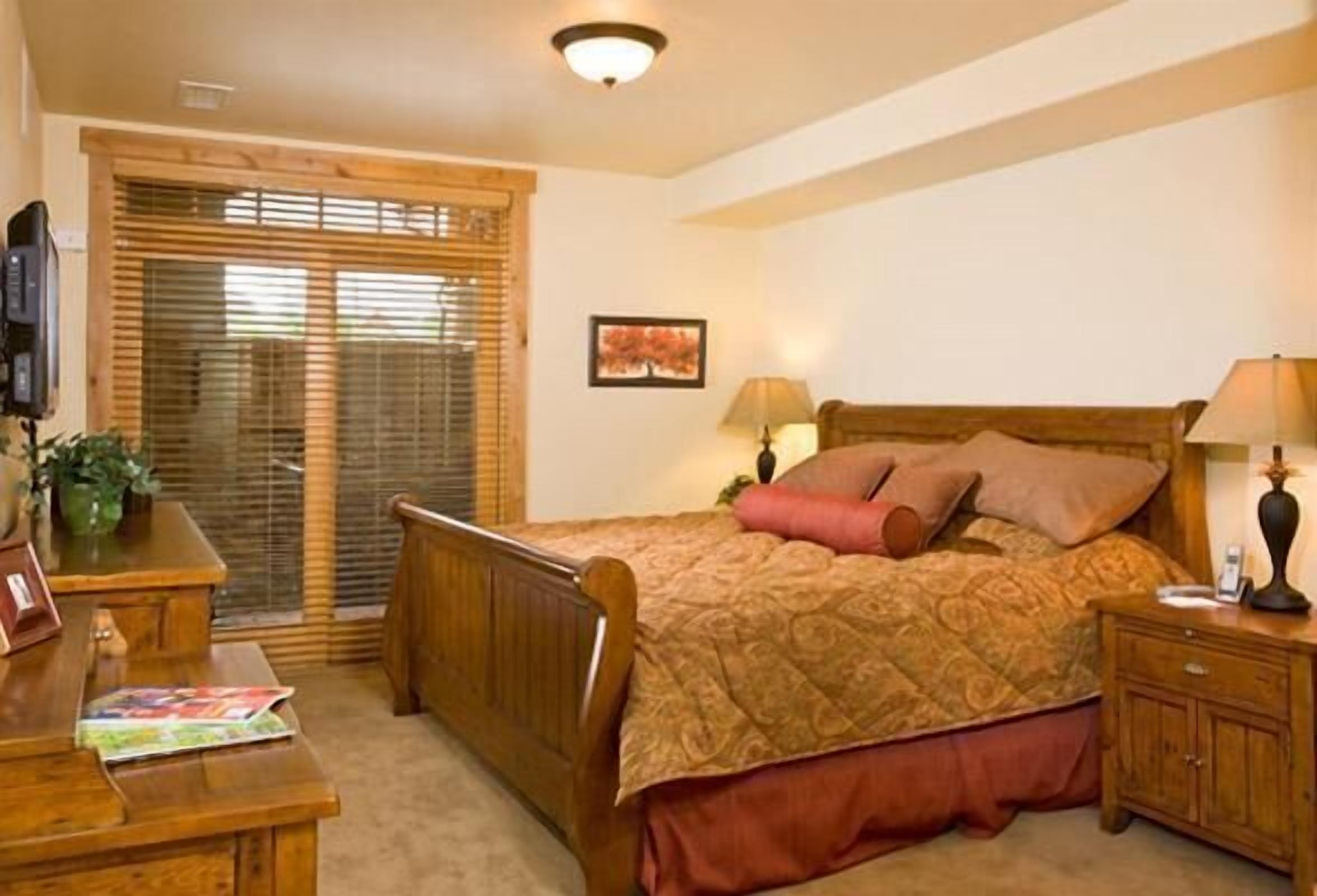 A bedroom with a wooden bed, a nightstand, a desk, and a window with blinds.