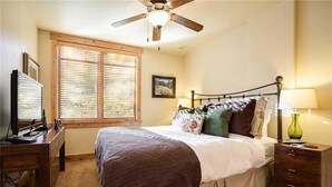 A bedroom with a bed, a desk, a ceiling fan, a window with blinds, and a nightstand with a lamp. - 6115 Bear Lodge, Trappeur's Crossing Apartment 1 (Steamboat Springs)