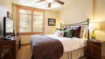 A bedroom with a bed, a desk, a ceiling fan, a window with blinds, and a nightstand with a lamp.