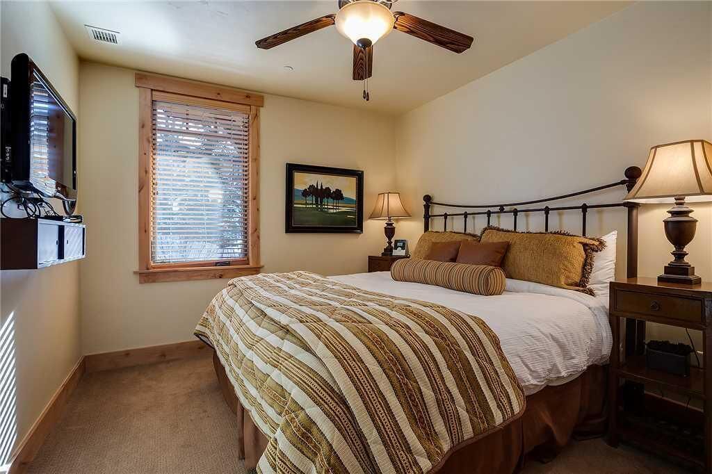 A bedroom with a bed, a ceiling fan, a TV, a window with blinds, a bedside table with a lamp, and a framed picture on the wall.