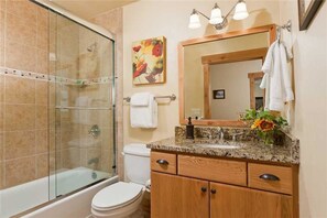 Un bagno con doccia in vetro, un water, un lavandino con piano in granito, armadietti in legno e uno specchio con un asciugamano appeso.