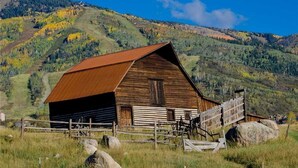 A rustic wooden barn with a metal roof, surrounded by a wooden fence and large rocks, set against a backdrop of rolling hills and autumn foliage. - 218 Storm Meadows Club B Apartment 1 (Steamboat Springs)