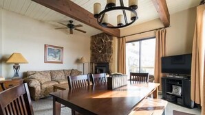 Un comedor con una mesa de madera, sillas y una chimenea de piedra. Hay un televisor y un ventilador de techo en la habitación.