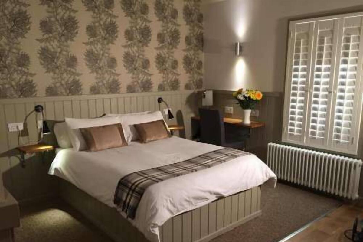 Quarto com cama, mesa de cabeceira, escrivaninha, cadeira, radiador e janela com persianas.