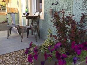 An outdoor seating area with two metal chairs and a table, a hanging metal bucket, and a planter with purple flowers. - Irvine Bay Studios (Irvine)