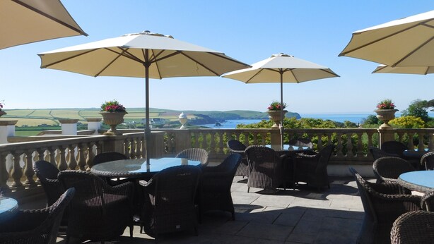 An outdoor patio with wicker furniture and large umbrellas, overlooking a scenic coastal landscape. - Thurlestone Hotel (Kingsbridge)