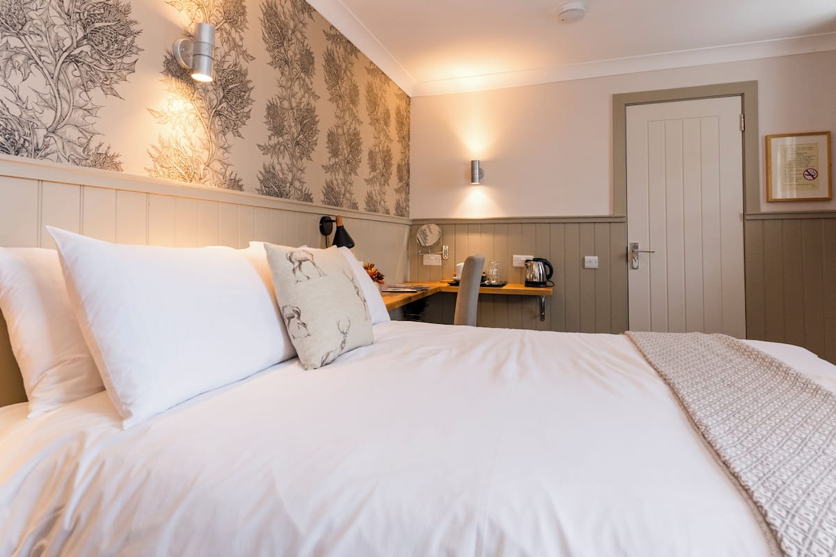 Quarto de hotel com uma cama grande, uma mesa, uma cadeira, uma luminária na parede e uma porta.