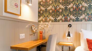 A room with a wooden desk, a chair, a bed with white pillows, a wall-mounted lamp, and a floral wallpaper.