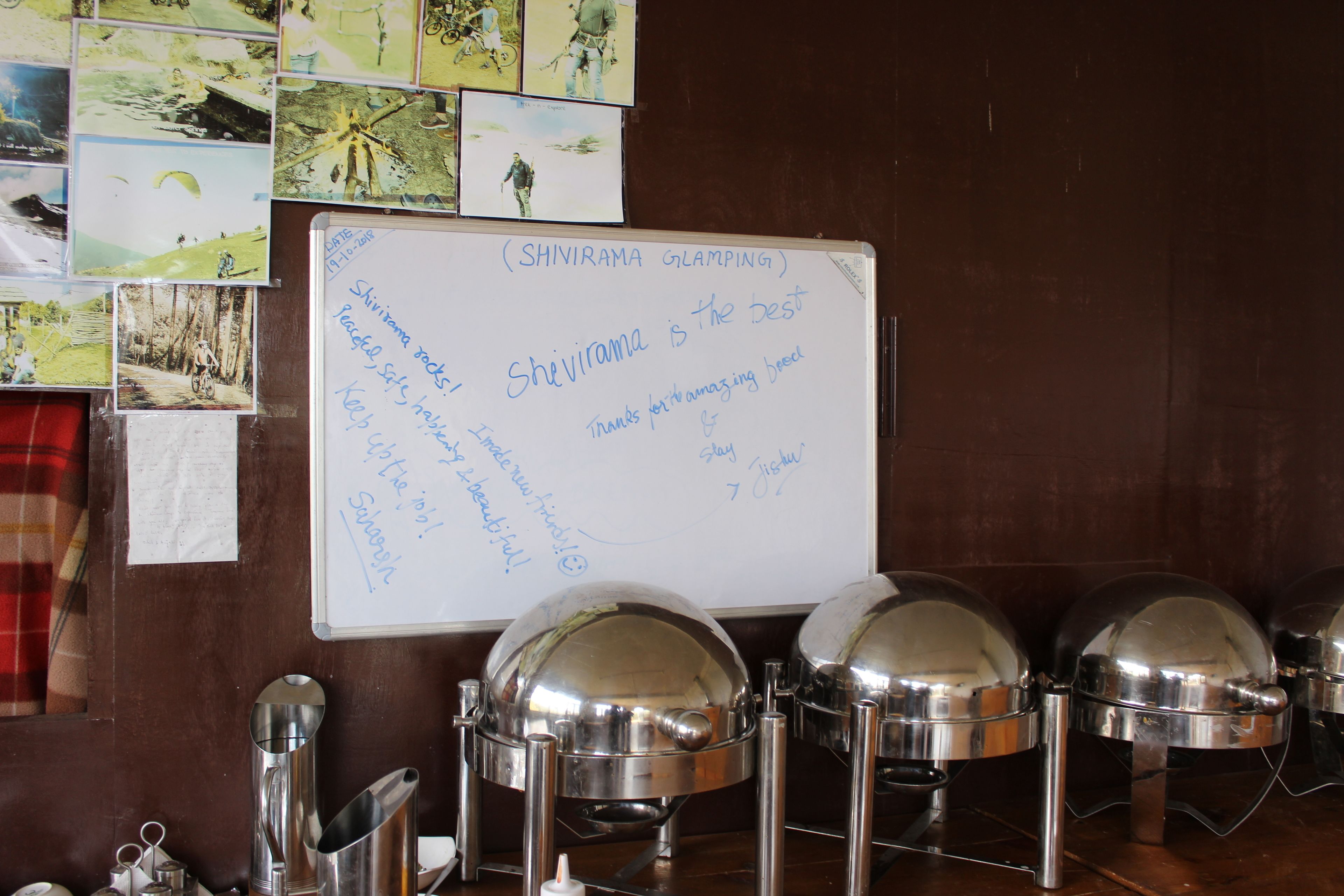 A whiteboard with handwritten notes and three covered food dispensers on a wooden table.
