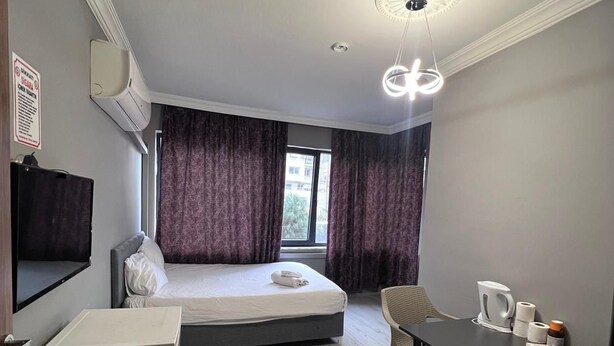 A compact hotel room with a bed, a small desk, a chair, a TV, and a window with curtains. - Gurleyen Apart Otel (Izmir)