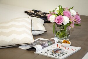 Un lit avec des oreillers, un magazine, un vase de fleurs et un saladier rempli de fruits.