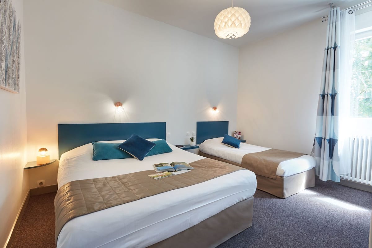 Une chambre d’hôtel avec deux lits, une grande fenêtre avec des rideaux, un luminaire fixé au mur et une petite table de chevet avec une lampe.