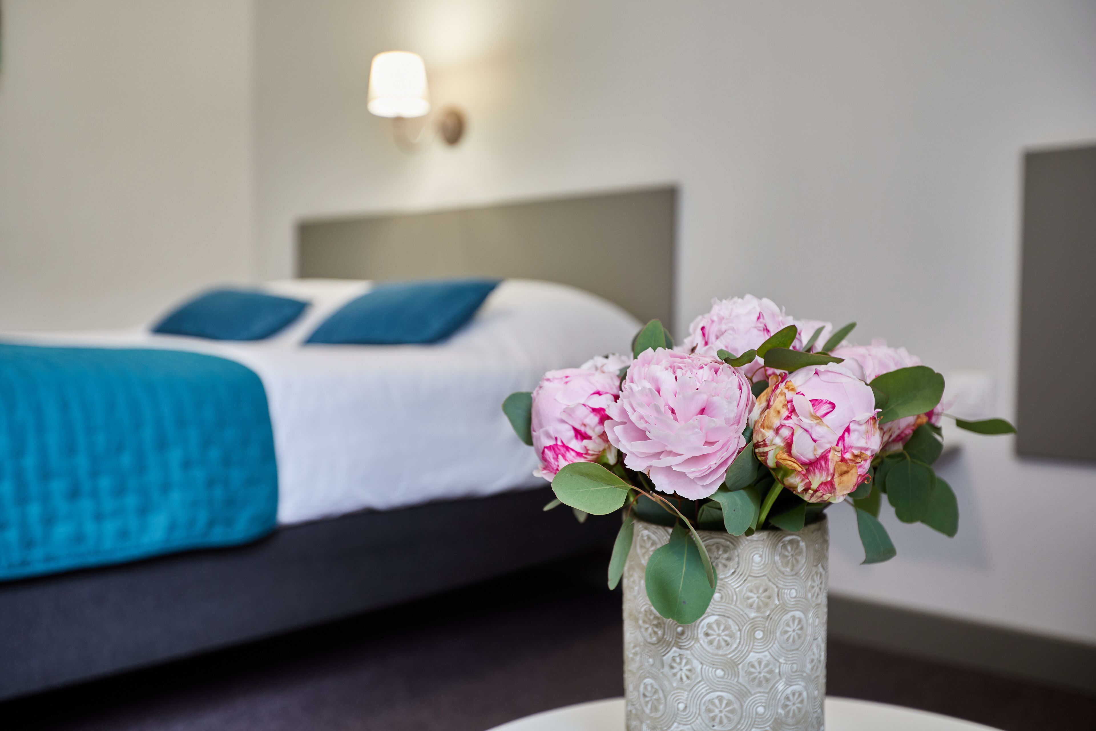 A neatly made bed with a blue blanket and pillows, a vase with pink flowers, and a wall-mounted light fixture.