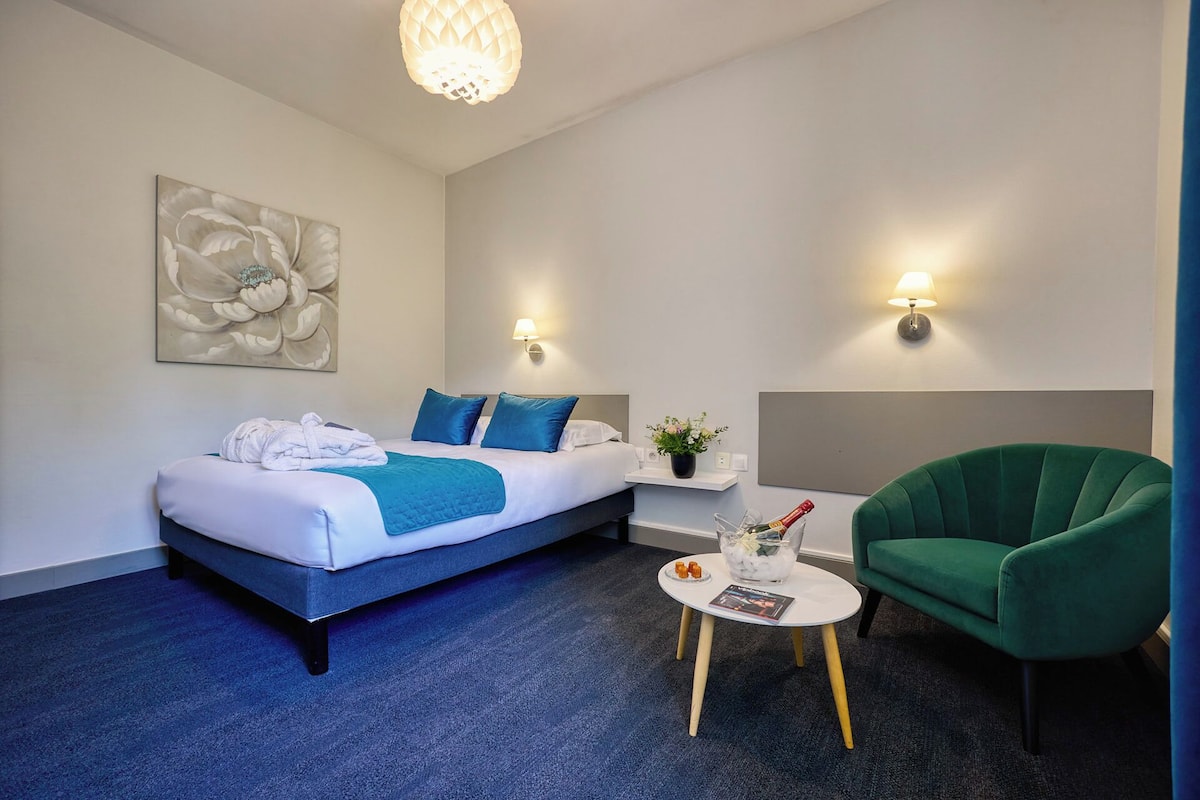 Une chambre d’hôtel avec un lit, un fauteuil vert, une petite table avec des en-cas et une lampe fixée au mur.