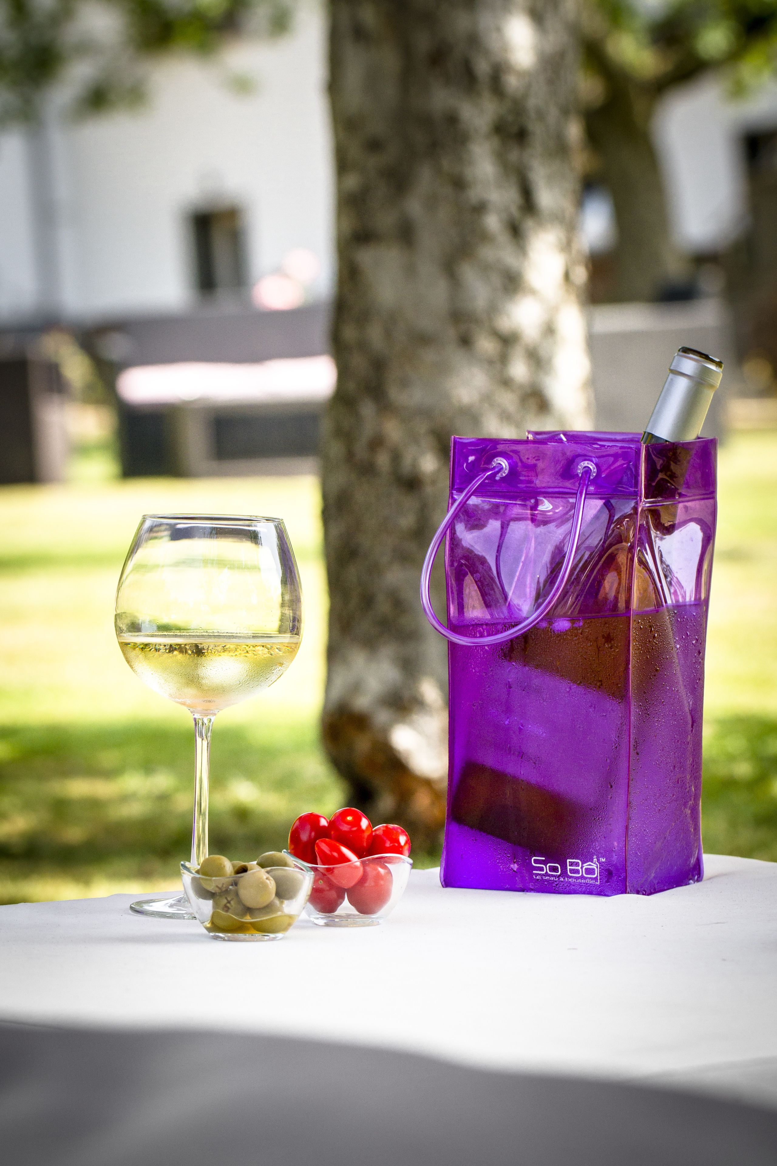 Un verre de vin blanc, un sac violet et quelques olives et tomates cerises sur une table à l’extérieur.