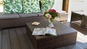 Un canapé en osier agrémenté de coussins à motifs de feuilles vertes, une table en osier avec un magazine et un vase rempli de fleurs.